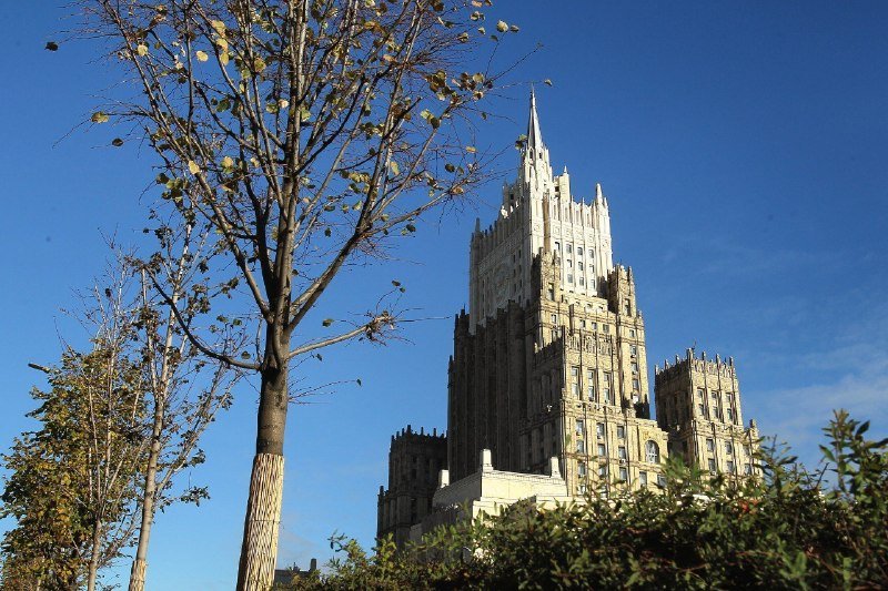A tall, ornate stone building with Gothic-style spires rises against a clear blue sky, framed by a leafless tree in the foreground.