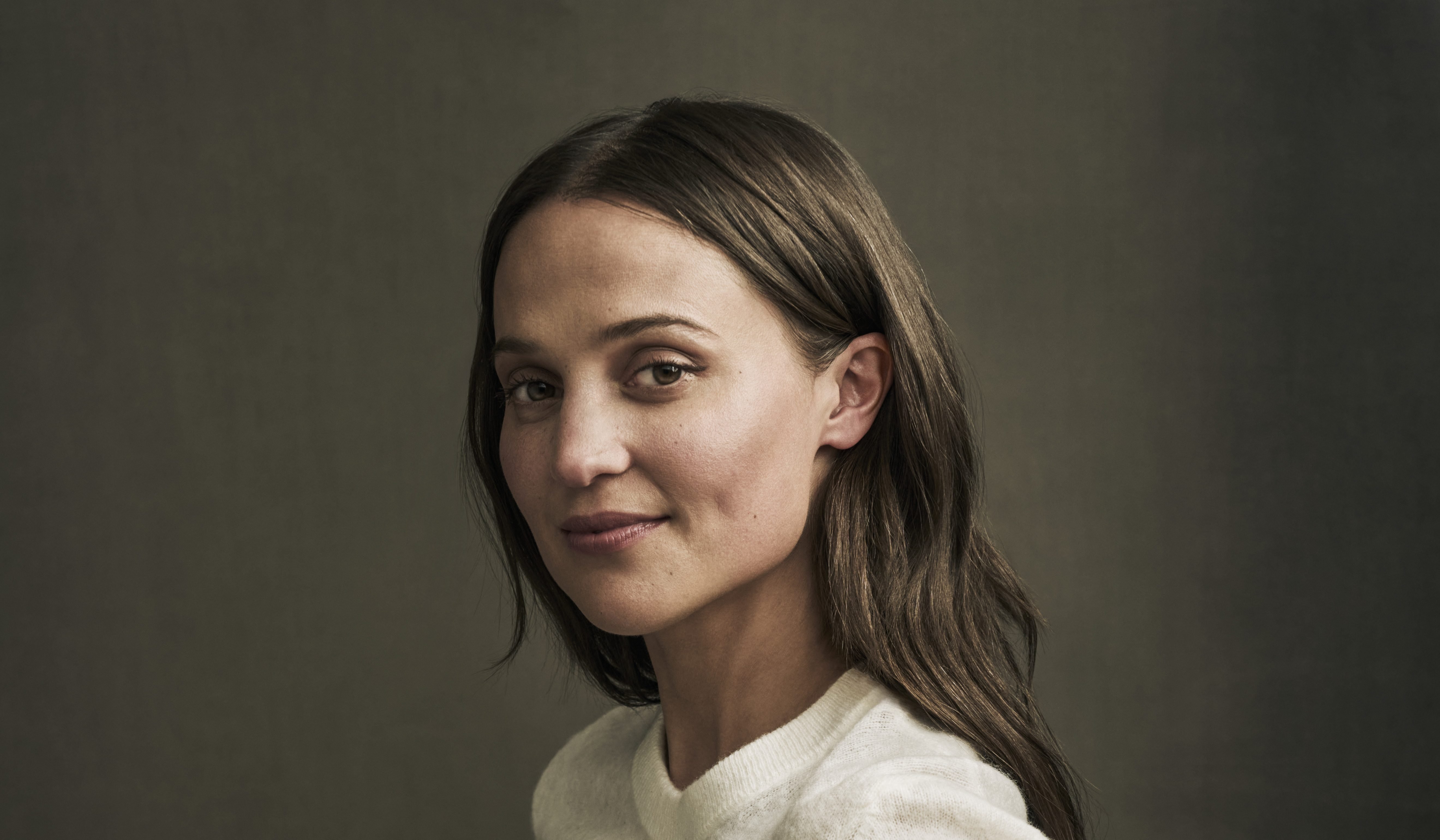 A woman with long brown hair wearing a cream-colored sweater poses for a portrait against a muted olive-green backdrop.