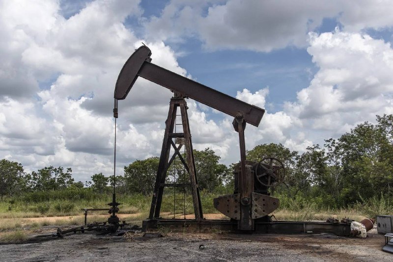 A rusted pumpjack stands in a grassy field under a partly cloudy sky, surrounded by trees.