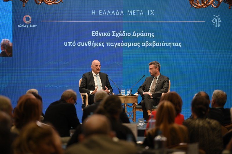 Two men in suits sit on stage at a podium with microphones, addressing a seated audience in a room with a blue backdrop displaying Greek text.