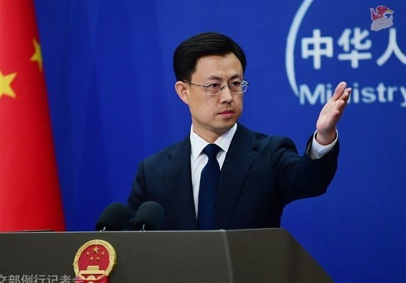 A bespectacled man in a dark suit gestures with one raised hand while standing at a podium bearing an emblem, with a Chinese flag and a blue backdrop with partial English text reading "Ministry" visible behind him.