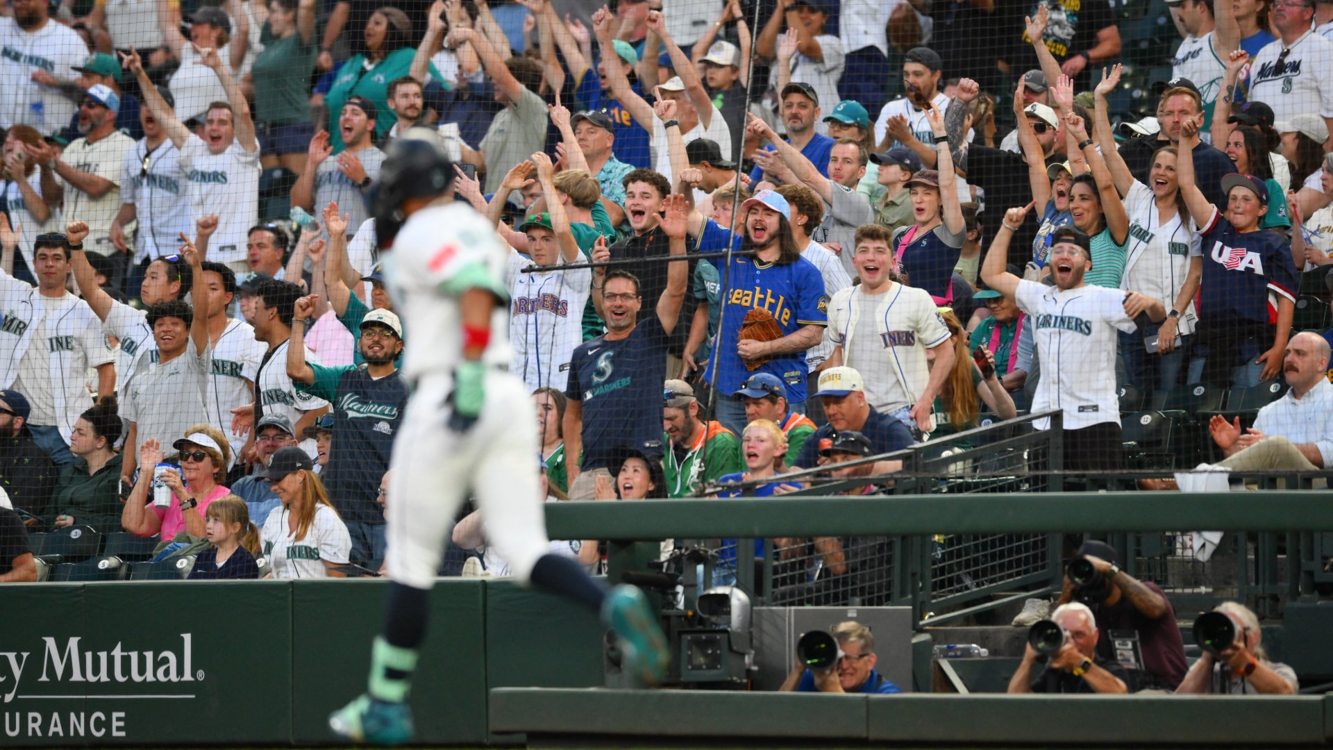 A baseball player runs toward the dugout as cheering fans in Mariners and Seattle jerseys raise their arms in celebration.