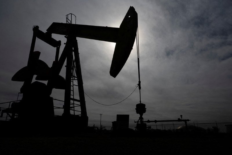 Silhouette of a pumpjack and an oil derrick stand against a cloudy sky with the sun breaking through behind them.