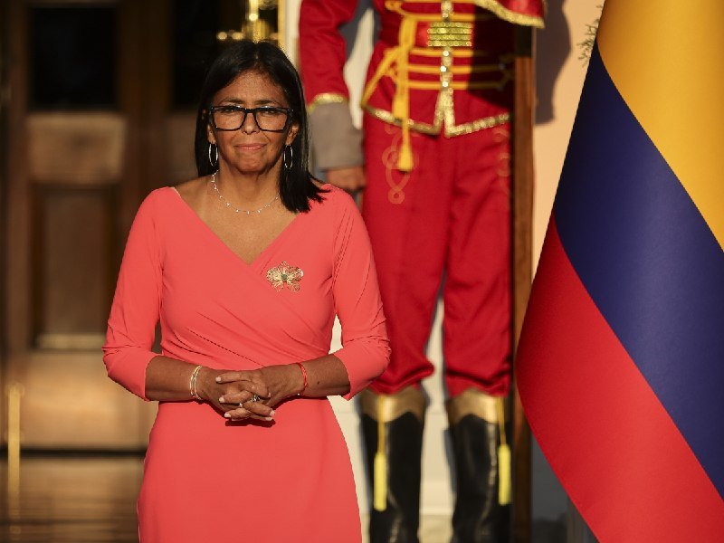 A woman in a coral dress and glasses stands beside a guard in a red ceremonial uniform and a yellow, blue, and red flag.