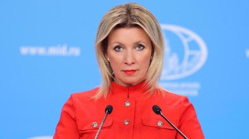 A blonde woman in a red high-collared jacket speaks at a podium with two microphones against a blue backdrop featuring a partial globe logo and web address.