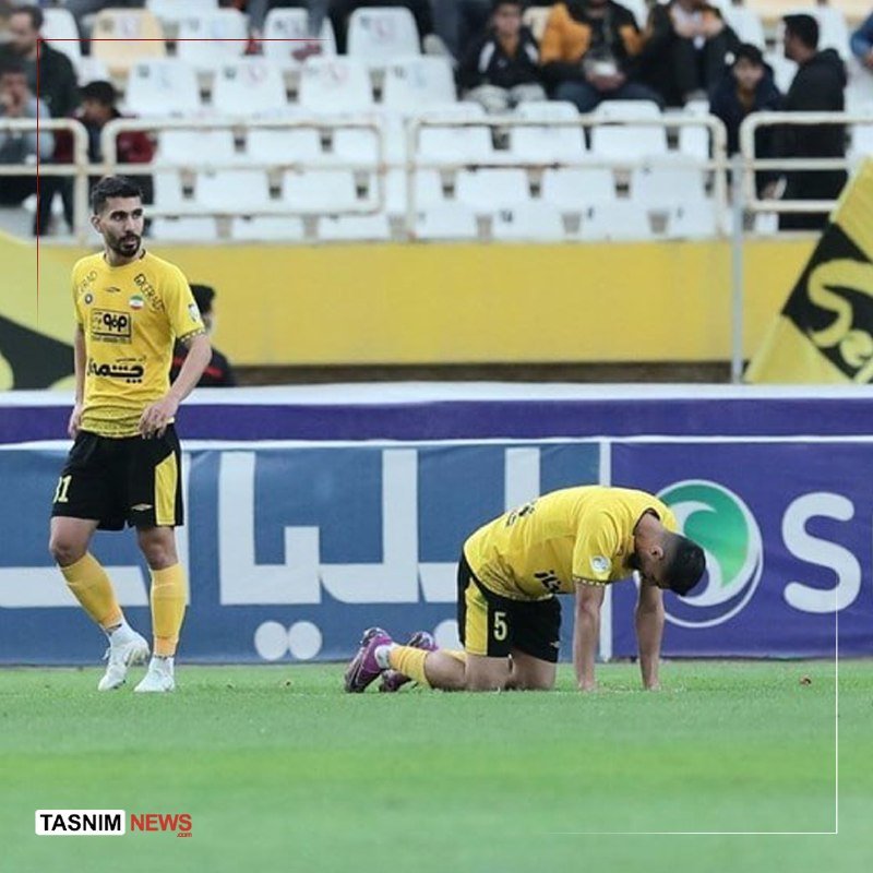 Two soccer players in yellow and black uniforms appear dejected on the field, with one standing and the other kneeling, while spectators sit in the stands behind them.