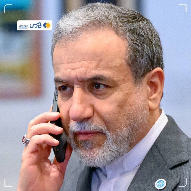 A gray-haired, bearded man in a dark suit holds a smartphone to his ear, with a small logo visible in the upper left corner.