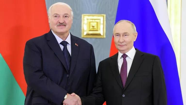 Two men in dark suits shake hands in front of a red-and-green flag and a white-blue-red flag, standing before a gray wall with a gold-framed decoration.