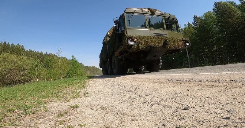 A large, mud-splattered military truck drives along a paved road bordered by grass and trees under a clear blue sky.