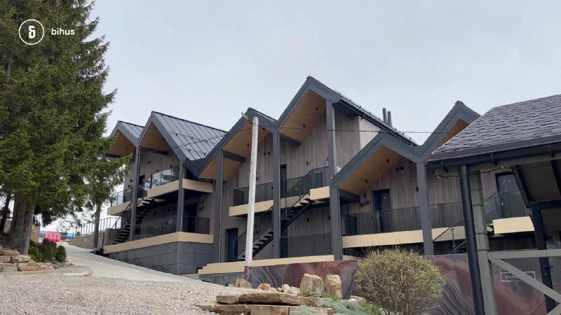 A row of modern, multi-story wooden chalets with steep triangular roofs and glass balconies stands along a gravel driveway bordered by evergreen trees and a metal decorative fence.