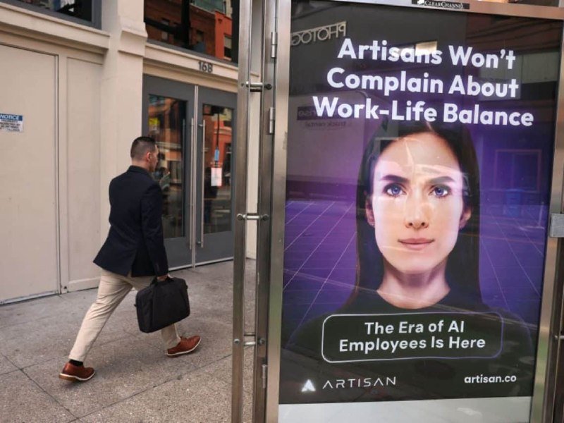 A man in a blazer carrying a briefcase walks past a large illuminated Artisan advertisement reading "Artisans Won't Complain About Work-Life Balance" and "The Era of AI Employees Is Here."