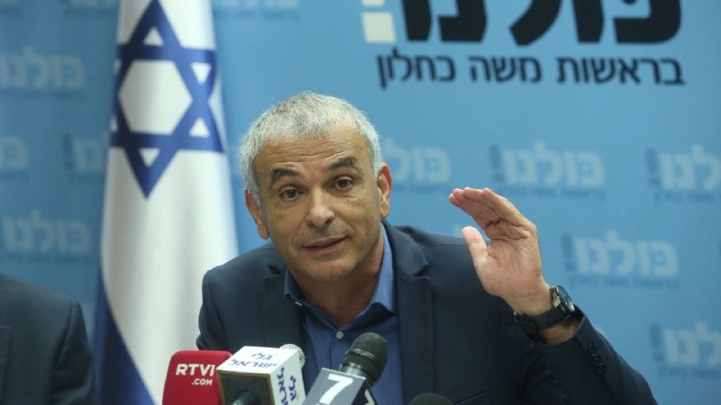 A gray-haired man in a dark suit gestures while speaking at microphones in front of an Israeli flag and a blue backdrop with Hebrew text.