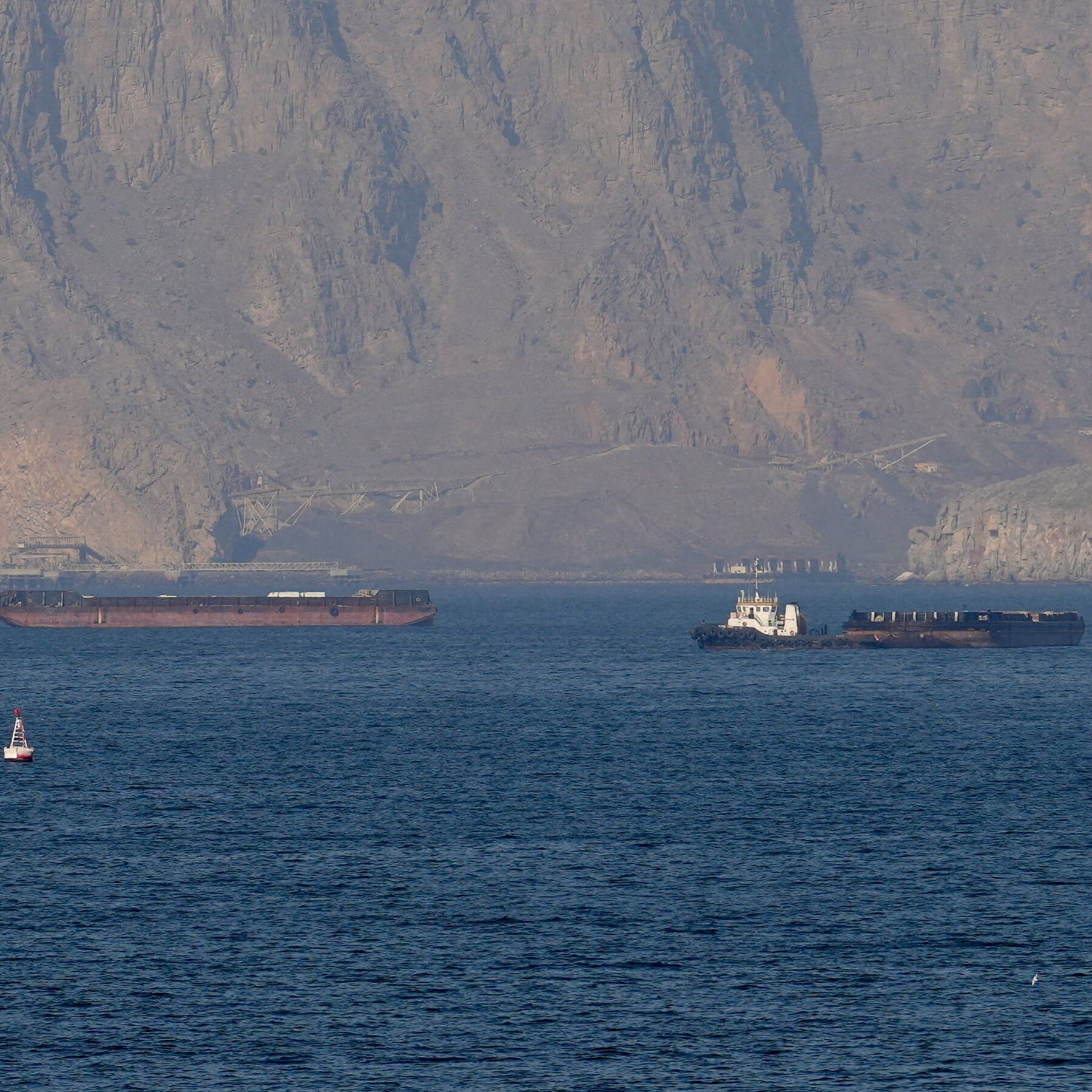 A tugboat pushes a barge loaded with dark material across a body of water, with a red cargo vessel and a small buoy also visible against a steep, rocky mountain backdrop.