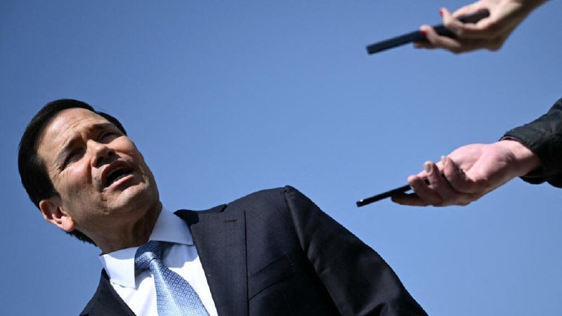 A dark-haired man in a suit and tie speaks while looking upward, as two hands holding recording devices extend toward him against a clear blue sky.