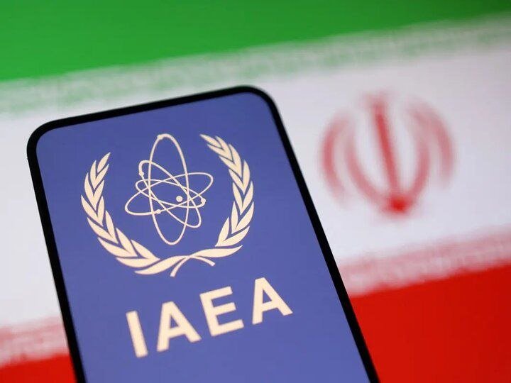 A smartphone displays the IAEA logo featuring an atom encircled by laurel wreaths, positioned in front of a blurred flag with green, white, and red stripes.