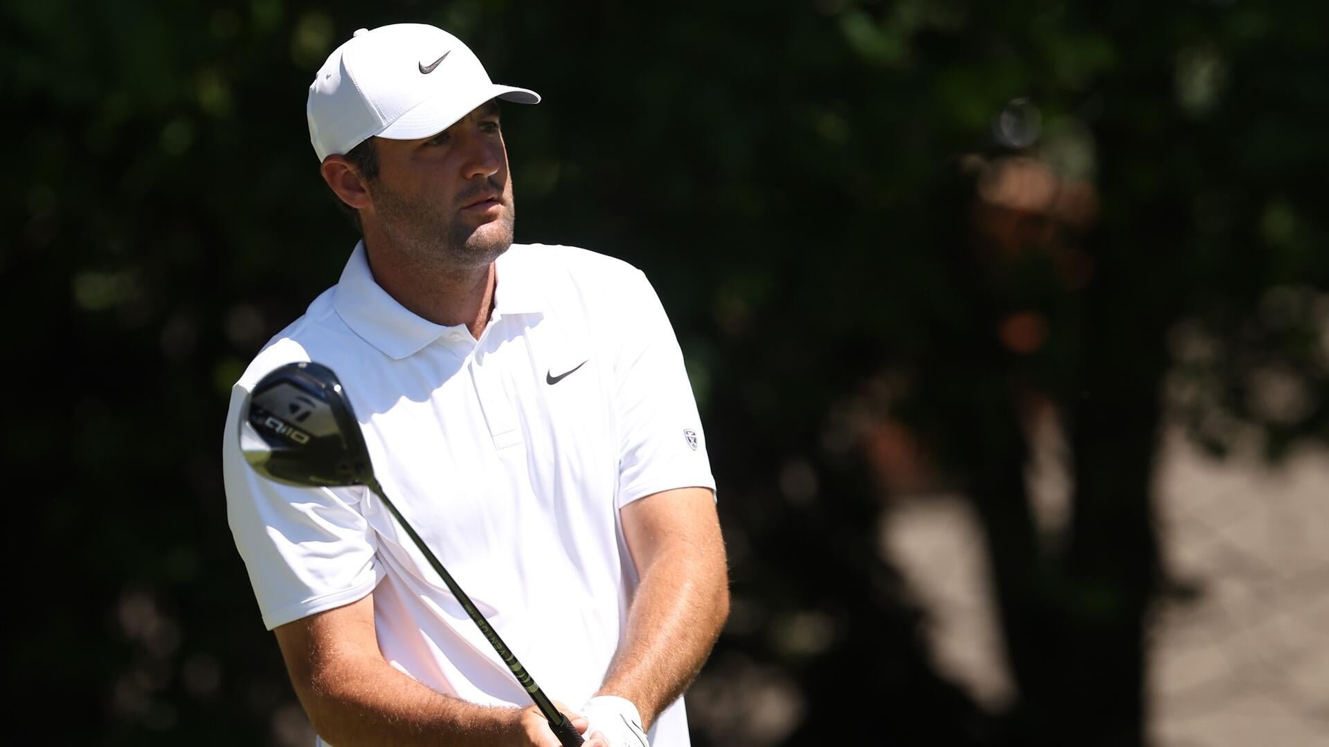 A man in a white Nike cap and shirt holds a golf club, watching his shot during a round of golf.