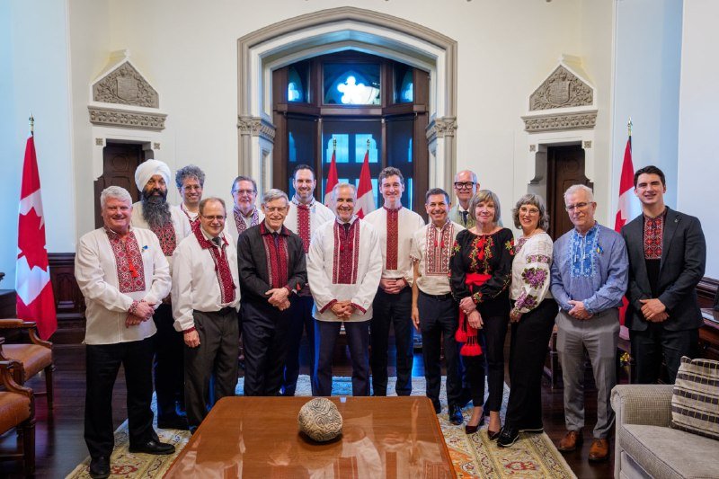 A group of people wearing embroidered shirts pose together indoors between two Canadian flags, with a wooden table in the foreground.
