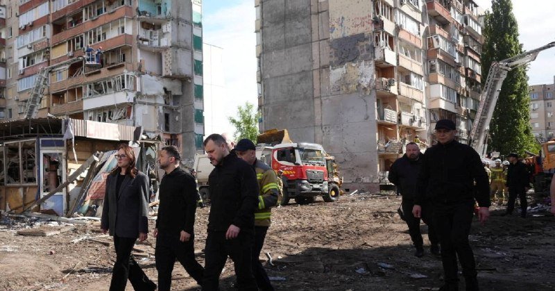 A group of people, including firefighters and men in dark clothing, walk through a debris-strewn area in front of a heavily damaged multi-story apartment building, with a fire truck and crane visible behind them.