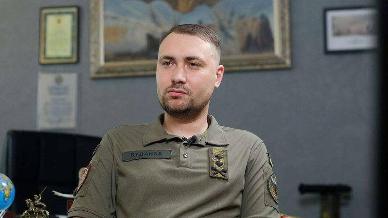 A bearded man wearing an olive uniform with a "БУДАНОВ" name patch sits in front of framed artwork and documents on a gray wall.