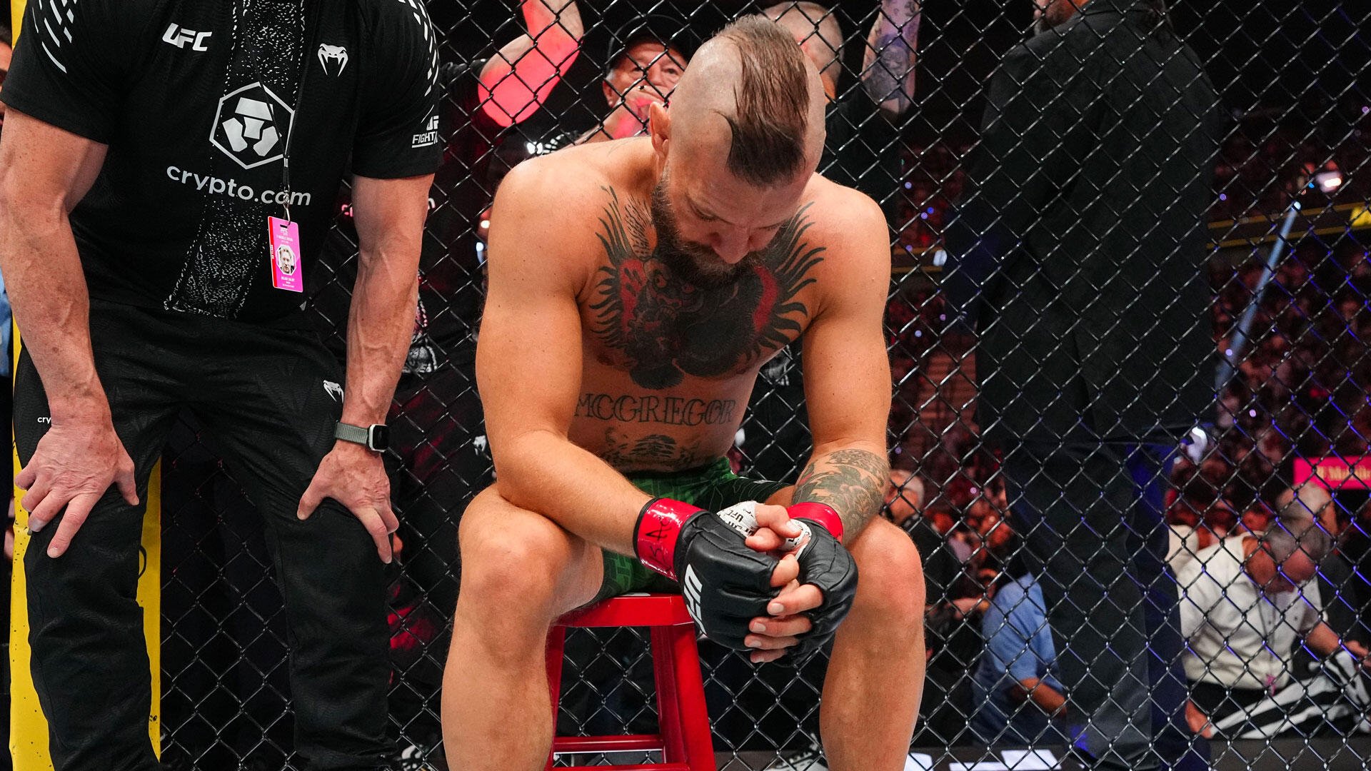 A shirtless, tattooed MMA fighter sits on a red stool inside a cage, head bowed and gloved hands clasped, beside an official in a black UFC/crypto.com shirt.
