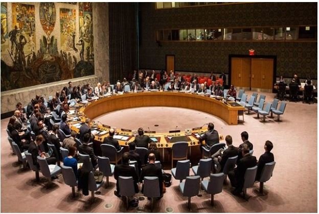 Officials seated around a large circular table in a formal council chamber with a mural on the wall.