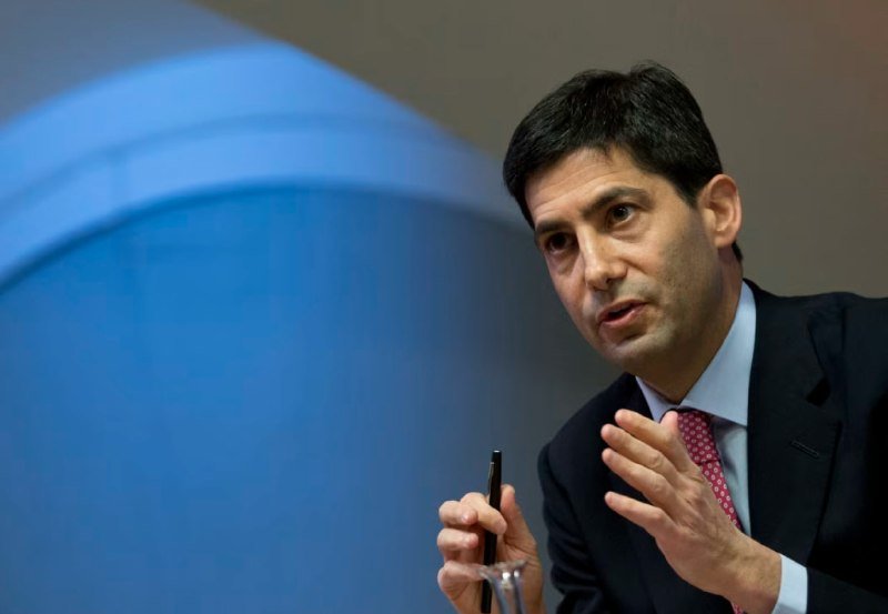A dark-haired man in a dark suit, light blue shirt, and red patterned tie gestures with his left hand while holding a pen, speaking against a curved blue backdrop.