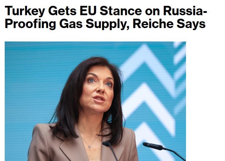 A dark-haired woman in a beige blazer speaks at a podium with microphones against a blue background featuring white geometric shapes.
