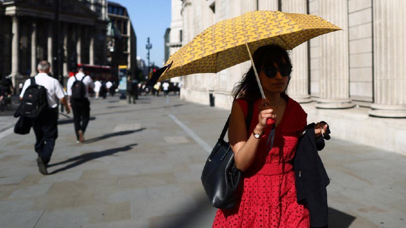 A woman in a red dress and sunglasses holds a yellow patterned umbrella on a sunny city street, carrying a black handbag and jacket.