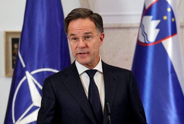A man in a dark suit and tie speaks at a podium, with a NATO flag and a Slovenian flag visible behind him.