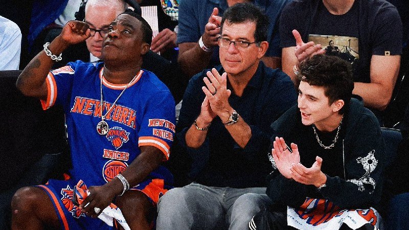 Three men sit courtside, clapping; one wears a blue New York Knicks jersey, another a dark polo with a watch, and the third a black sweater with a silver chain.