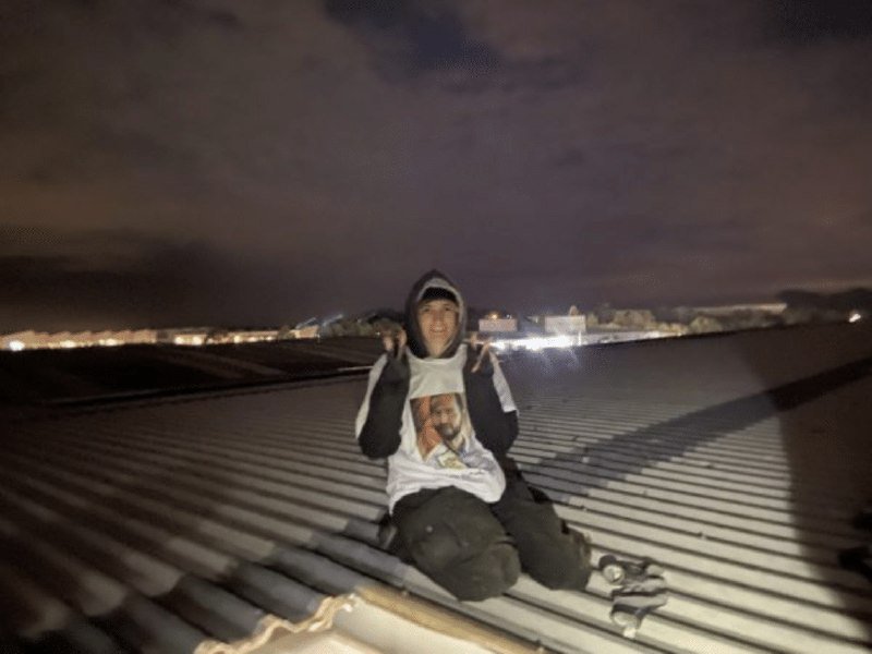 A person in a hooded sweatshirt sits on a corrugated rooftop at night, smiling toward the camera, with distant city lights visible on the horizon.