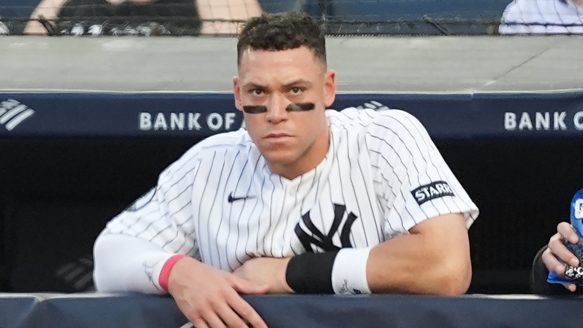 A baseball player in a white pinstriped Yankees jersey leans on the dugout railing with a serious expression, eye black visible under his eyes.
