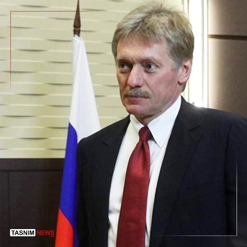 A gray-haired, mustachioed man in a dark suit and red tie stands beside a Russian flag, with a "Tasnim News" watermark visible.