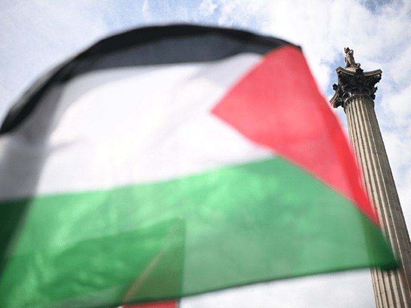 A Palestinian flag waves in the foreground with a tall stone column topped by a statue visible against a partly cloudy sky in the background.