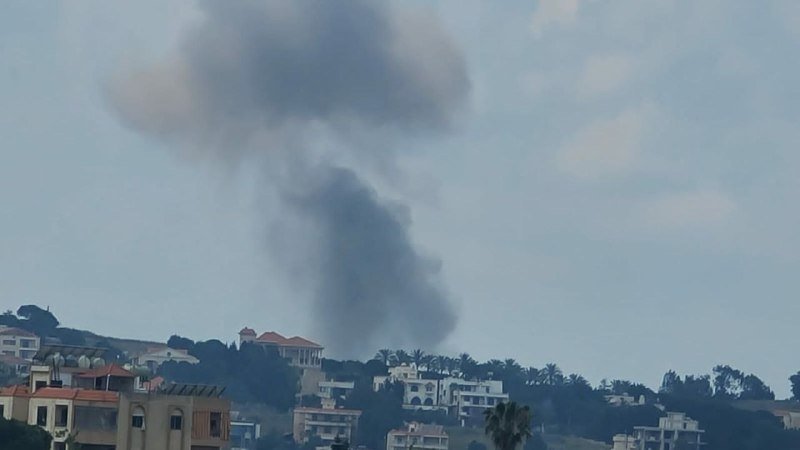 A large plume of dark gray smoke rises into the sky above a hillside residential area with buildings and palm trees in the foreground.