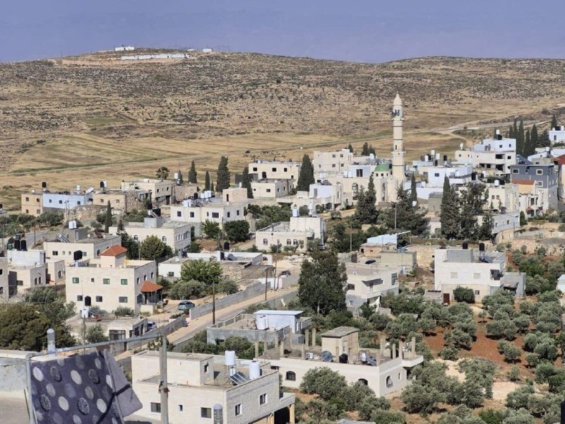 White and beige buildings cluster in a village on a hillside, featuring a tall minaret, scattered trees, and arid mountains in the background.