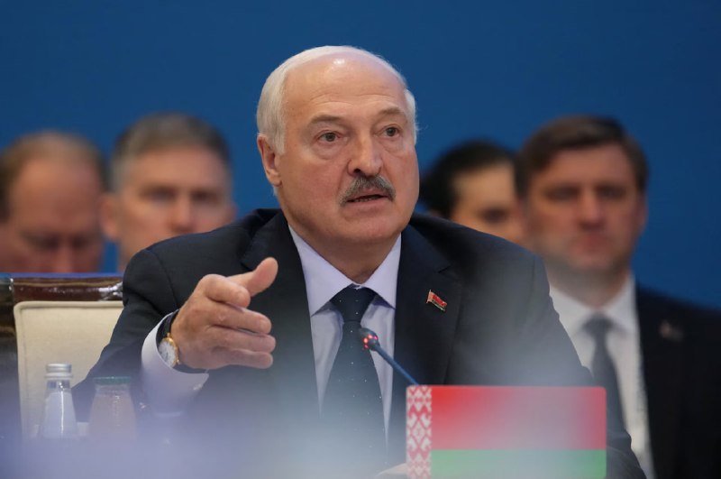 A white-haired man in a dark suit gestures while speaking into a microphone, with a Belarusian flag visible in the foreground and other people seated behind him.