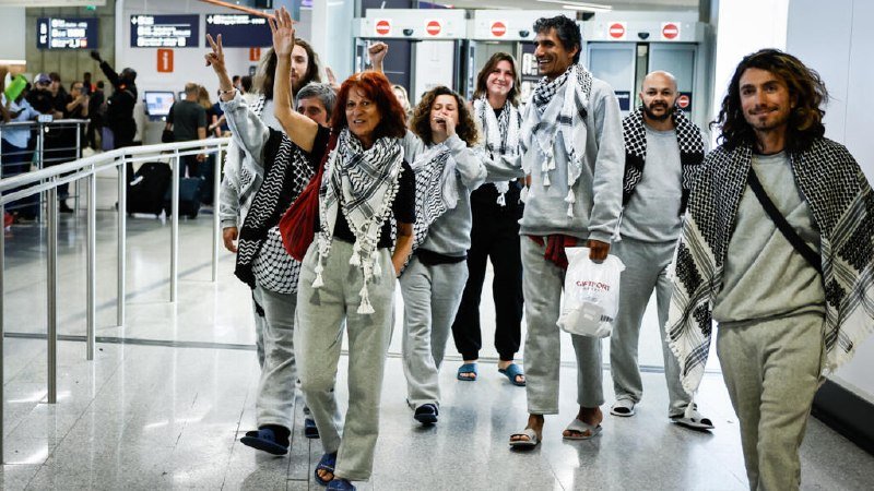 A group of people wearing matching gray sweatshirts and black-and-white keffiyeh scarves walk through an airport terminal, some raising their arms while one carries a tote bag.