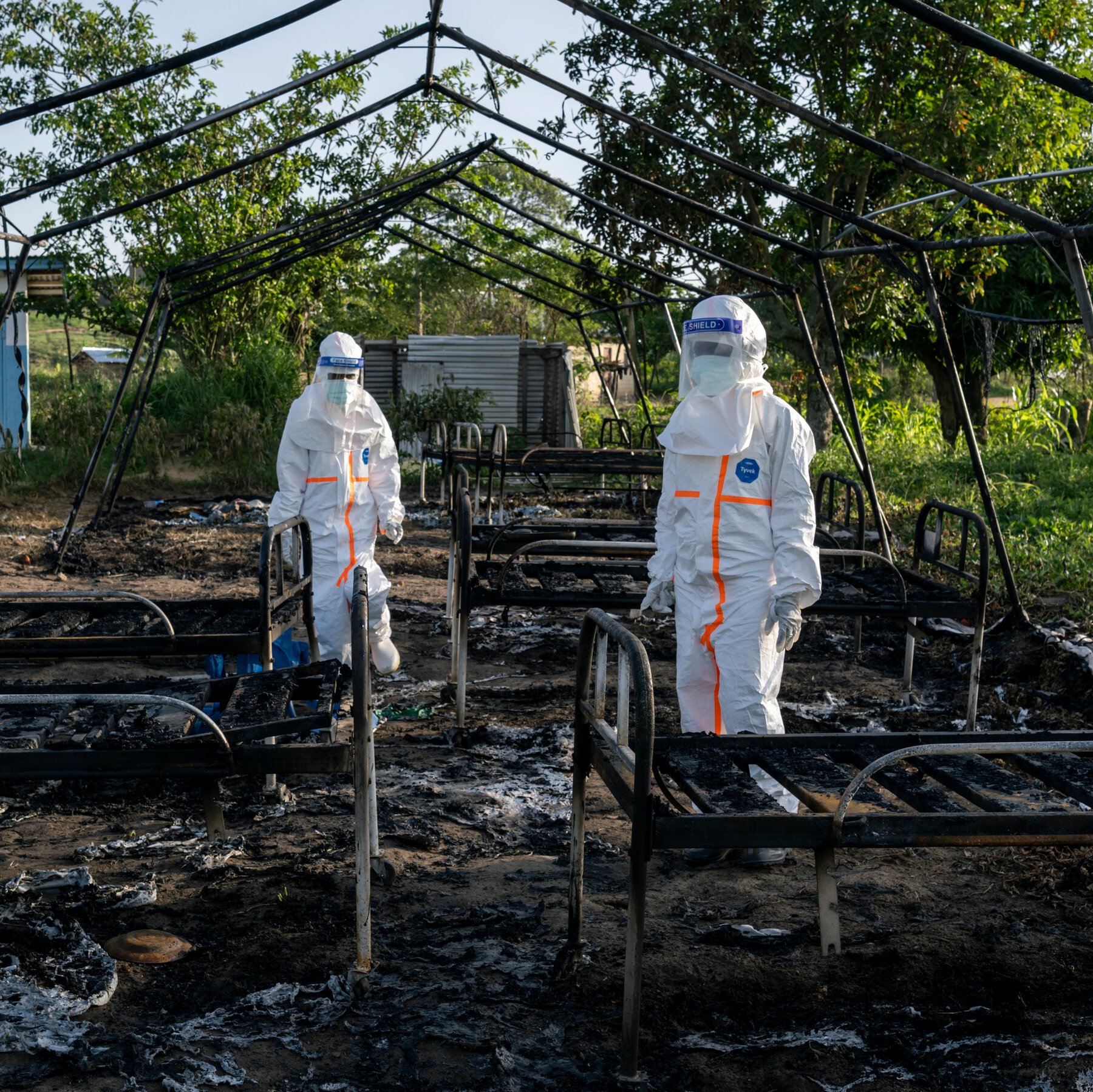 Two people in white hazmat suits with face shields stand among charred, blackened metal bed frames inside the burnt-out frame of a large structure.