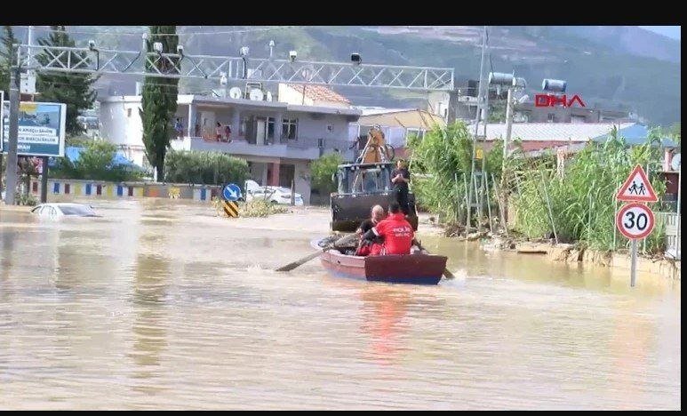 Rescue workers in a small boat and a tractor navigate a flooded street with submerged vehicles, buildings, and road signs visible in the background.