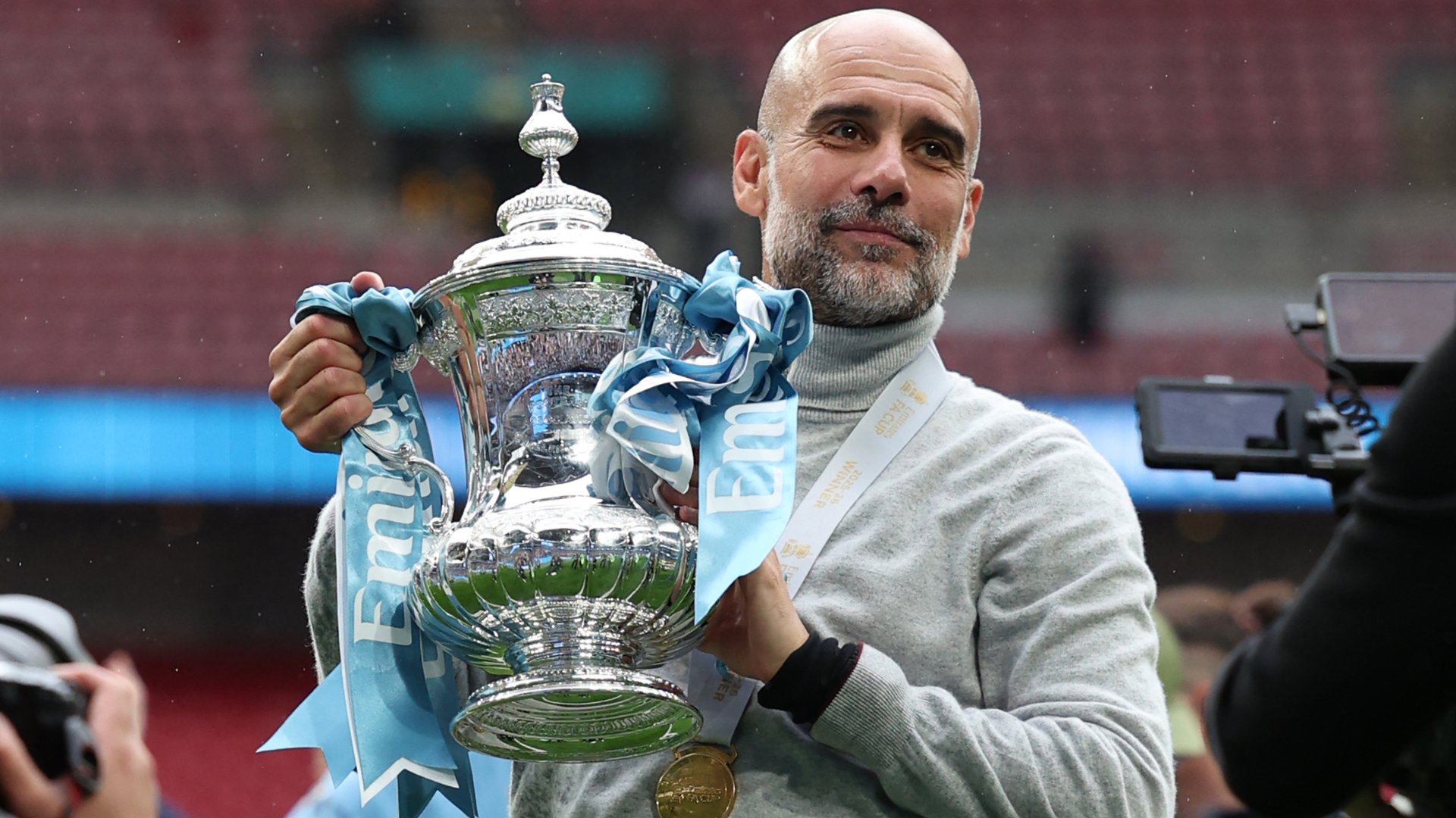 A bald man in a gray turtleneck holds a large silver trophy adorned with blue ribbons inside a stadium, with a medal around his neck.