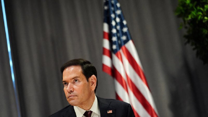 A man in a dark suit with an American flag pin sits before gray curtains and an unfurled U.S. flag.