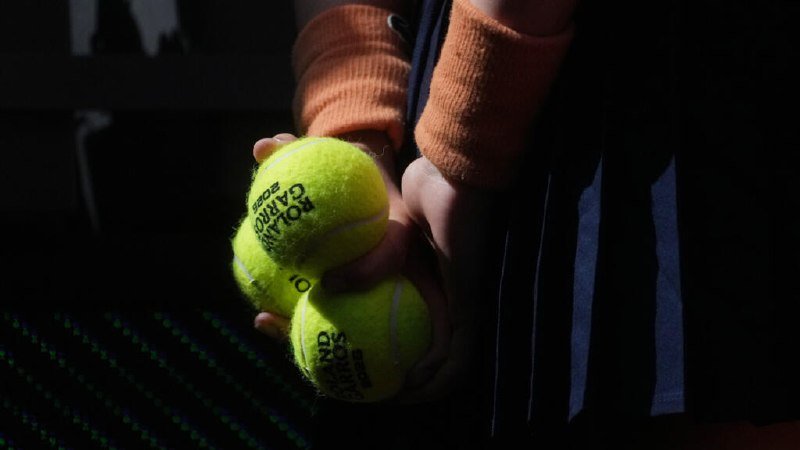 A person wearing an orange sweater holds three yellow Roland-Garros tennis balls in their hands against a dark background.