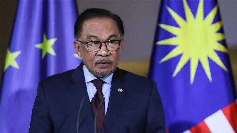 A bespectacled man in a dark suit and burgundy tie speaks at a podium, with a blue EU flag and a Malaysian flag visible behind him.