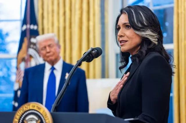 A woman in a black blazer speaks at a podium with a presidential seal, her hand on her chest, while an older man in a blue suit stands behind her.