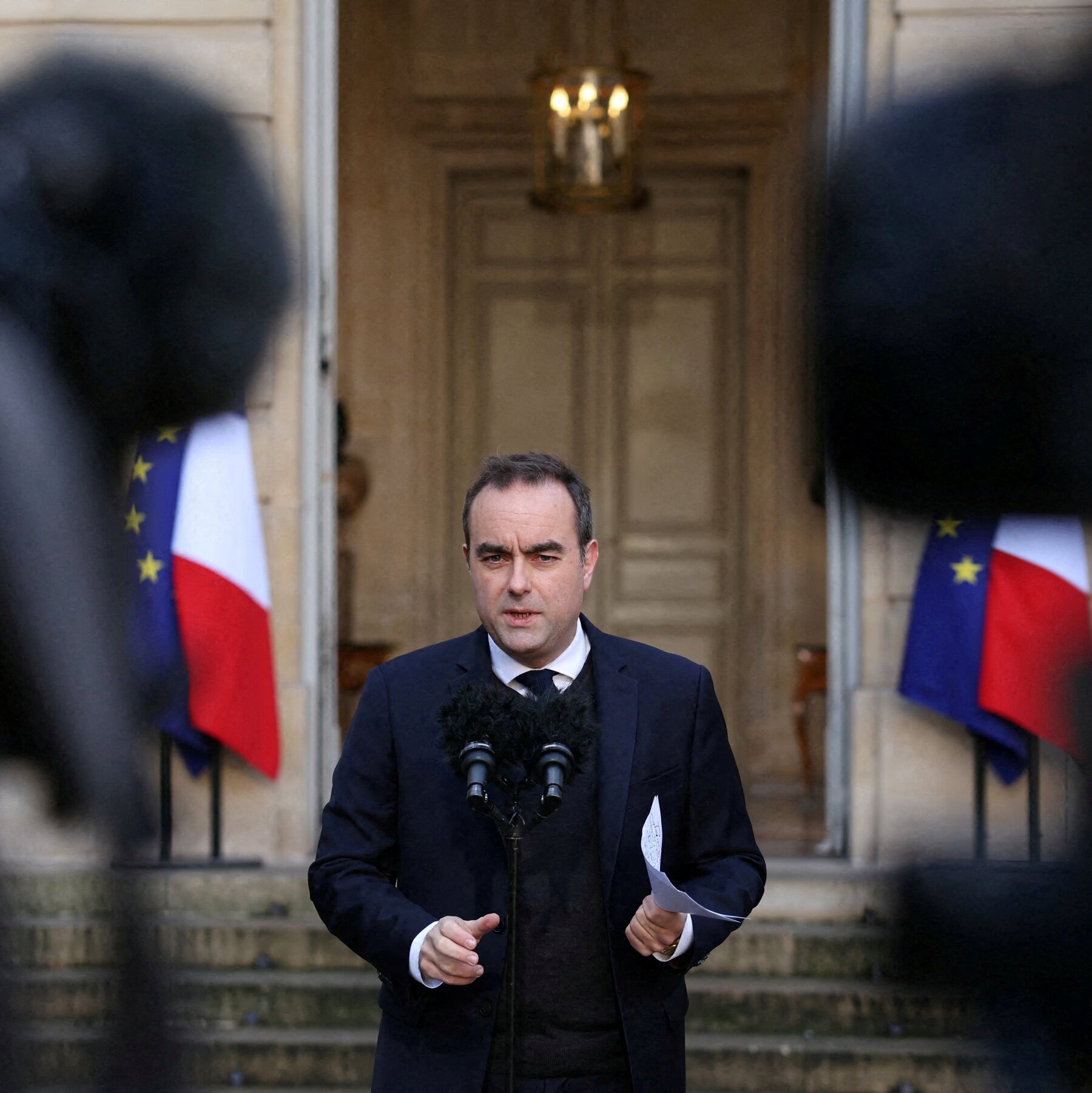 A man in a dark suit and tie speaks at microphones outside a building entrance, holding papers, with French and EU flags displayed behind him.