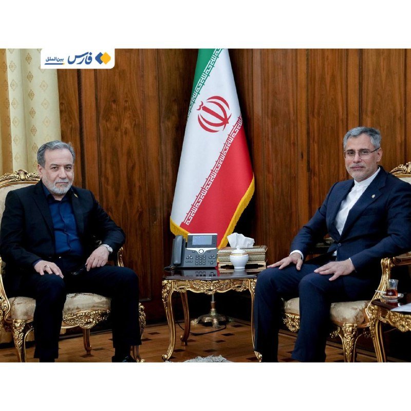 Two men in suits sit in ornate chairs facing the camera, with an Iranian flag displayed between them in a wood-paneled room.