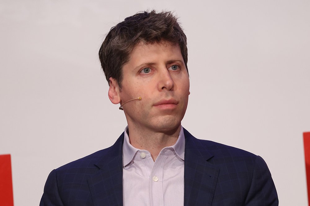 A man in a navy blazer and light button-up shirt with an earpiece speaks at a podium against a plain backdrop.