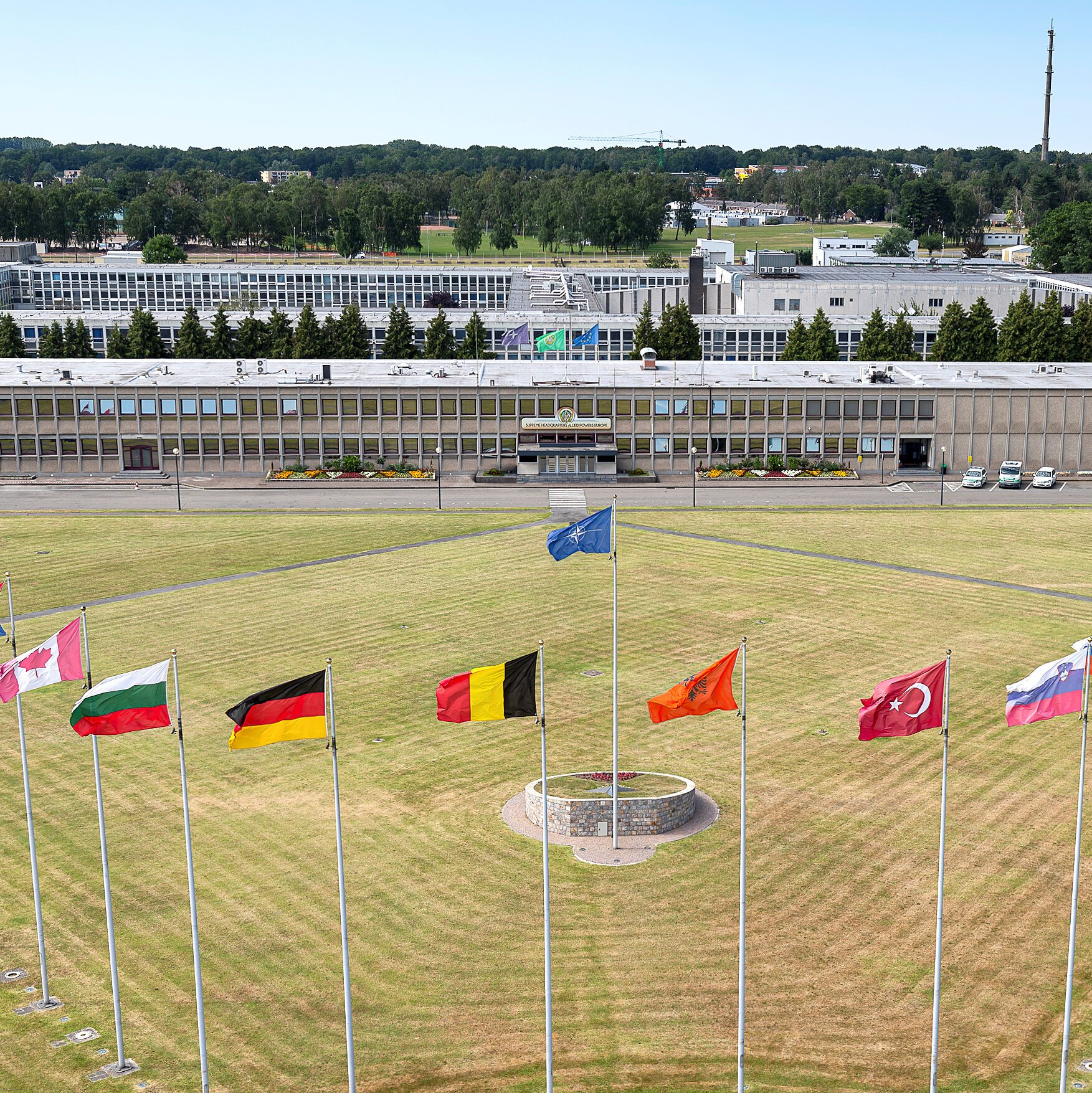 A large NATO headquarters building is shown behind a circular lawn displaying NATO, national, and other organizational flags on flagpoles, with a stone memorial at the center.