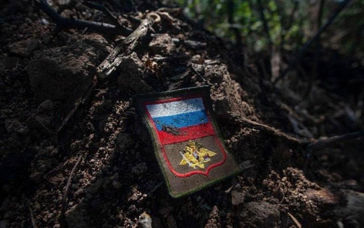 A mud-covered military patch featuring the Russian flag and an emblem rests on disturbed earth in a wooded area.
